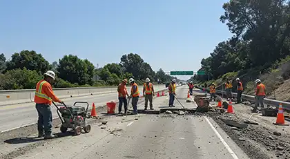 Road workers in orange vests repairing a highway lane surrounded by cones and debris