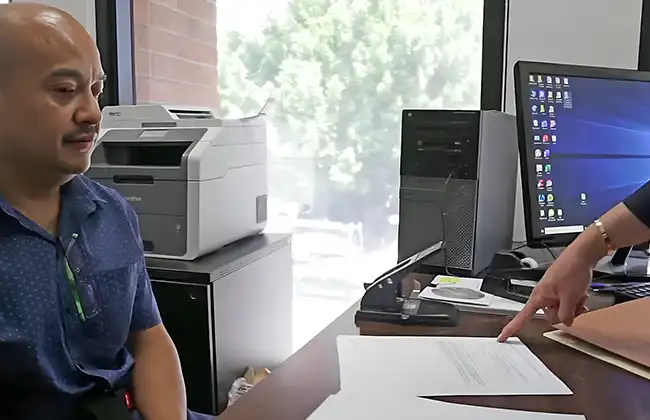 man sits in an office while another person points to documents on a desk