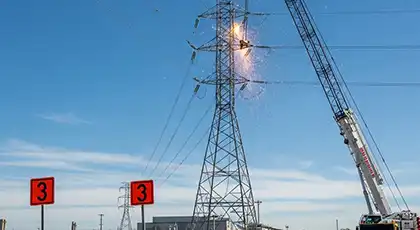 Grúas trabajando cerca de una torre eléctrica alta, con chispas que salen del contacto con la línea eléctrica