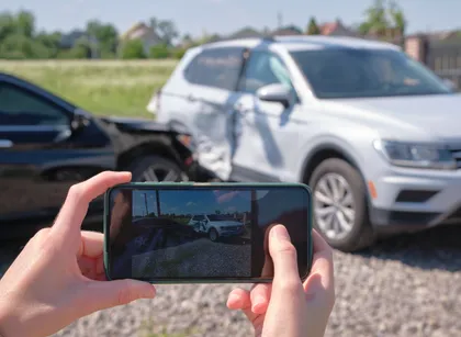 Person records auto accident damage on smartphone with blurred crash scene and dented SUV in background