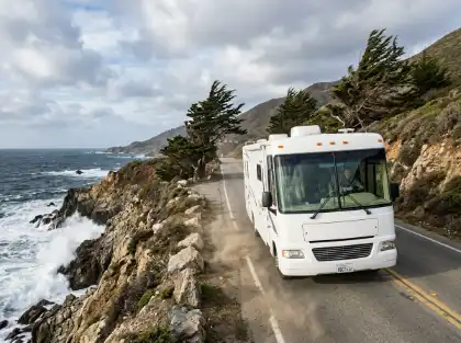 Una camioneta blanca que recorre una sinuosa carretera costera de California junto a acantilados rocosos y las olas del mar