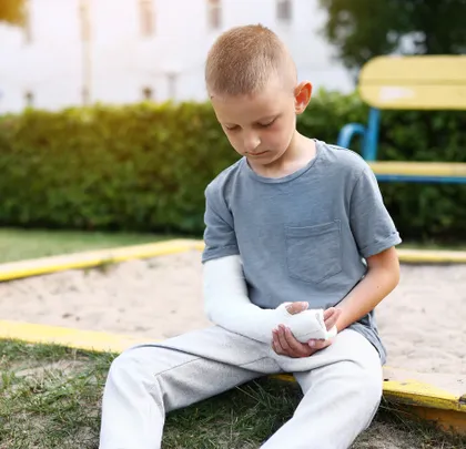 Un niño pequeño con un brazo roto y una escayola blanca, sentado junto al arenero de un parque infantil al aire libre