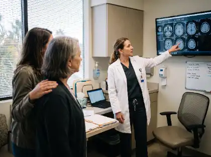 Una doctora que señala unas imágenes de una resonancia magnética cerebral en una pantalla mientras se las explica a dos mujeres en una consulta médica.